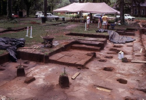 Archeological digsite. Tents and cards are in the background while tools and wheelbarrows are in foreground.