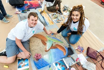 Photo of a teenage boy and teenage girl smiling with a chalk drawing on a sidewalk.